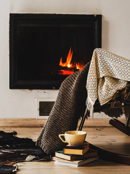 Rocking chair with knit rug, books and cup of tea or coffee before fireplace Stock Photo