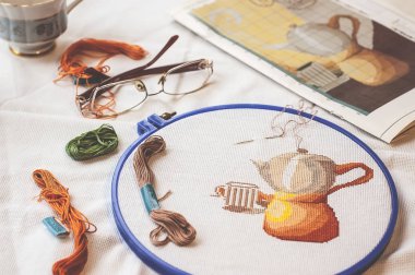 Cross-stitch set : hoop with embroidered pattern, glasses, scheme, scissors, canvas and colorful yarn. Selective focus. Freelance, hobby, handmade home decor concept clipart