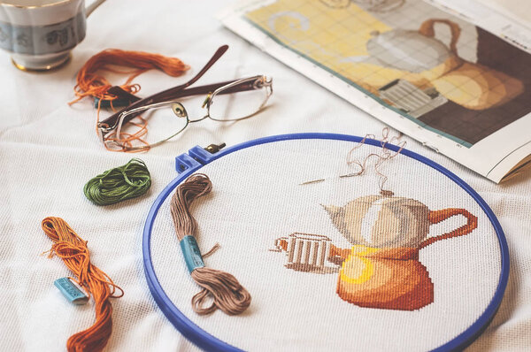 Cross-stitch set : hoop with embroidered pattern, glasses, scheme, scissors, canvas and colorful yarn. Selective focus. Freelance, hobby, handmade home decor concept