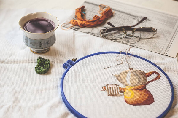 Cross-stitch set : hoop with embroidered pattern, glasses, scheme, scissors, canvas and colorful yarn. Selective focus. Freelance, hobby, handmade home decor concept