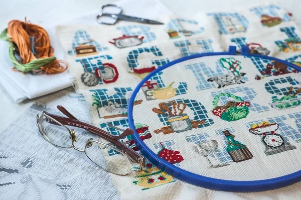 Cross-stitch set : hoop with embroidered pattern, glasses, scheme, scissors, canvas and colorful yarn. Selective focus. Freelance, hobby, handmade home decor concept — Stock Photo, Image