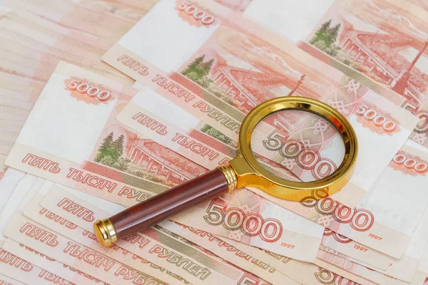 A magnifying glass with a gold handle is placed on top of Russian banknotes of five thousand rubles in large quantities