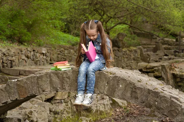 Little schoolgirl is reading a book in nature. Smart little pupi — Stock Photo, Image