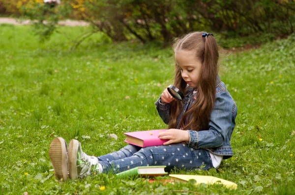 Una chica está leyendo un libro con una lupa. Inteligente poco p — Foto de Stock