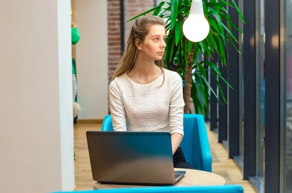 Retrato de bela mulher sorridente sentada em uma cadeira confortável em um café com laptop preto. Estudante bonito fazendo trabalho com laptop . — Fotografia de Stock