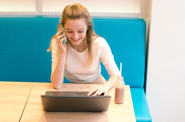 Ritratto di bella donna sorridente seduta su una comoda sedia in un caffè con computer portatile nero. Bella studente facendo il lavoro con il computer portatile e parlando al telefono . — Foto Stock