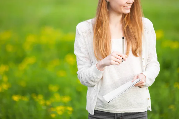 Young freckled meisje landbouwingenieur of bioloog in witte blouse staande in groene veld met notitieboekje en pen tijdens de oogst. — Stockfoto
