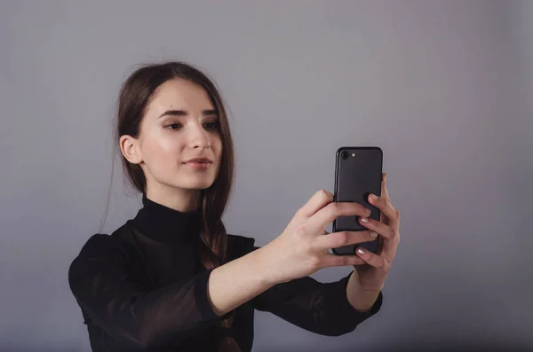 Potret seorang gadis muda yang menarik membuat foto selfie dengan smartphone terisolasi pada latar belakang abu-abu netral . — Stok Foto