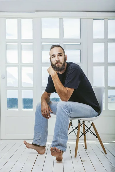 Young man in jeans with a beard a hipster sitting on a chair on