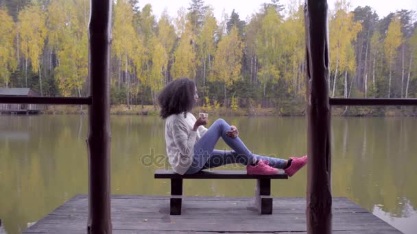 Красивая девушка сидит на скамейке и пьет горячий чай у озера — стоковое видео