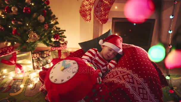 Kleine jongen met speelgoed klok viel in slaap tijdens het wachten voor Santa Claus in de buurt van de kerstboom met geschenken — Stockvideo