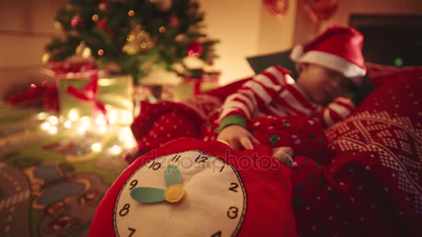 Маленький мальчик с игрушечными часами заснул в ожидании Санта Клауса возле рождественской елки с подарками — стоковое видео