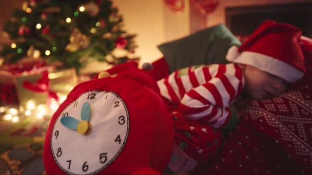 Close-up van speelgoed klok en het slapen van de kleine jongen droomt van Santa. Kerstboom met giften op de achtergrond — Stockvideo