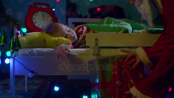 El niño duerme en su cuna en Navidad. Mamá lo trae presente vistiendo traje de Papá Noel y besándose en la mejilla — Vídeo de stock