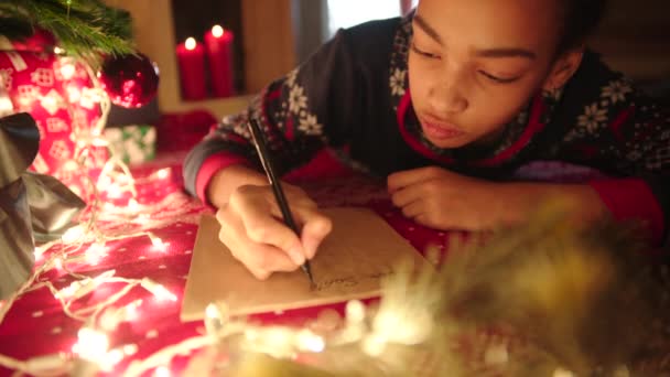 Teen girl is lying near decorated christmas tree and writing christmas letter to Santa Claus — Stock Video