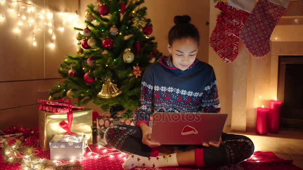 Mujer joven está hablando en línea con su familia usando el ordenador portátil en la noche de Año Nuevo. Ella está sentada cerca del árbol de Navidad decorado y la chimenea con calcetines de Navidad — Vídeo de stock