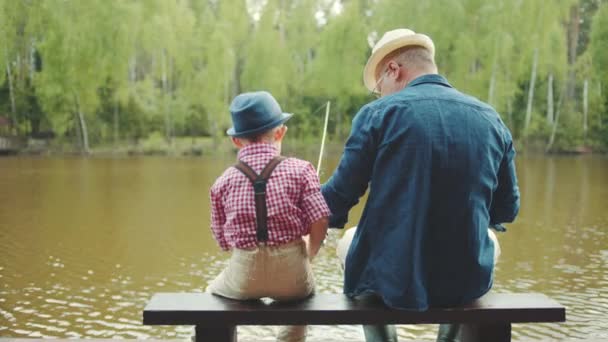 Velho e seu neto em chapéus de palha estão pescando no rio no verão. Eles estão segurando varas em suas mãos — Vídeo de Stock