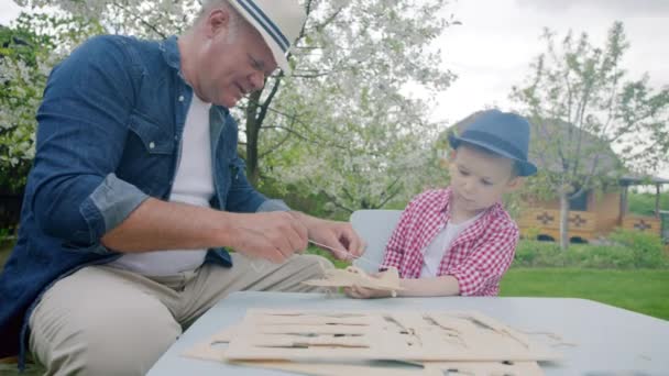 Grandpa is telling to his grandson how to make a wooden plane in the backyard — Stock Video