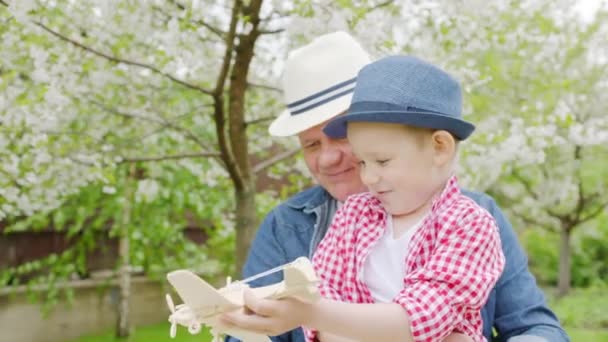 Jongen speelt houten vliegtuig met zijn opa in het voorjaar bloeiende tuin — Stockvideo