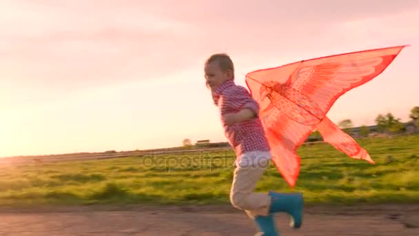 Мальчик бежит по проселочной дороге и улыбается с летящим воздушным змеем на закате — стоковое видео