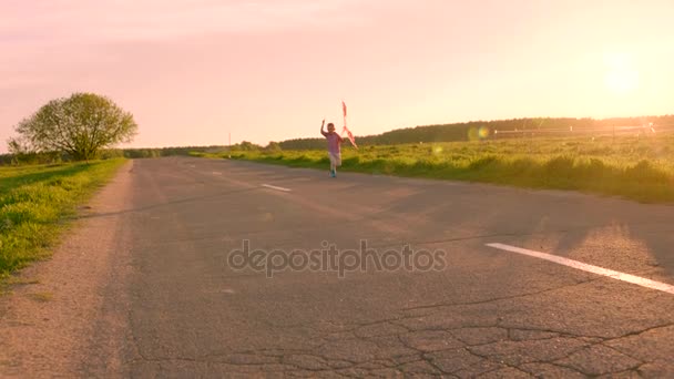 Niño corriendo por un camino rural y sonriendo con cometa voladora al atardecer — Vídeo de stock