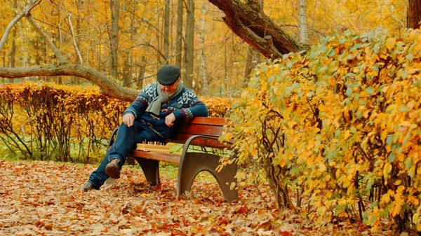Velho está jogando xadrez sozinho no banco no parque de outono — Fotografia de Stock