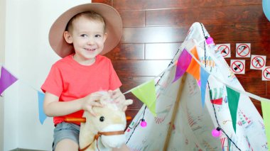 Kovboy şapkası giyen küçük mutlu çocuk oyun odası oyuncak ata biniyor