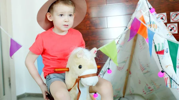 Kleiner glücklicher Junge mit Cowboyhut reitet auf einem Spielzeugpferd im Spielzimmer — Stockfoto