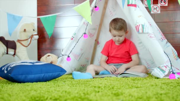 Pequeño niño está leyendo libro en su auto hecho wigwam en la sala de juegos — Vídeos de Stock