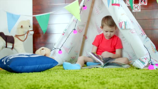 Pequeño niño está leyendo libro en su auto hecho wigwam en la sala de juegos — Vídeos de Stock