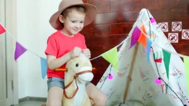 Kovboy şapkası giyen küçük mutlu çocuk oyun odası oyuncak ata biniyor