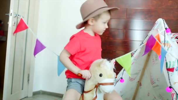 Menino feliz vestindo chapéu de cowboy está montando um cavalo de brinquedo na sala de jogos — Vídeo de Stock