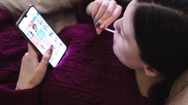 一个年轻姑娘吸着棒棒糖 看着智能手机 — 图库视频影像