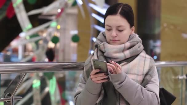 Dziewczyna patrzy na wyświetlacz smartfonów na tle kawiarni w centrum handlowym. Młoda atrakcyjna kobieta z telefonem w ręku w restauracji — Wideo stockowe