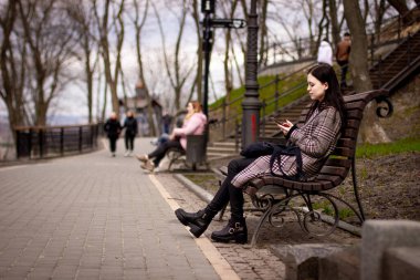 Parkta bankta oturan ve telefon kullanan esmer kadının yan görüntüsü.