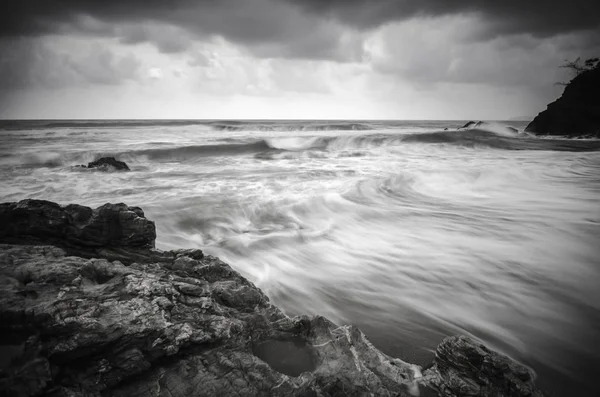 Flujo de ondas de imagen en blanco y negro en la playa arenosa y rocosa con nubes oscuras y dramáticas . — Foto de Stock