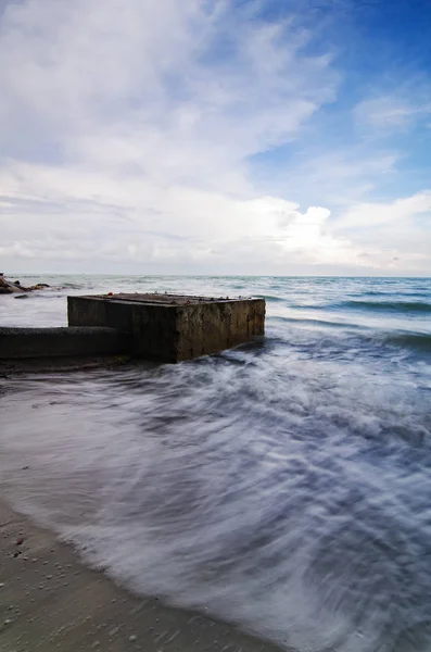 Weiche weiße Welle trifft Betonstruktur am Strand. verschwommenes Bild durch Langzeitbelichtung. Weiche und dramatische weiße Wolken mit blauem Himmel — Stockfoto