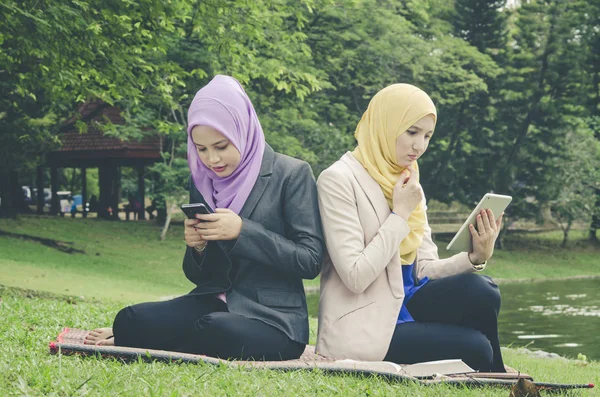 Joven muslimah sentado en una hierba verde en el parque y leer el libro electrónico en su teléfono móvil —  Fotos de Stock