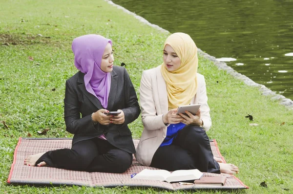 Joven muslimah sentado en una hierba verde en el parque y leer el libro electrónico en su teléfono móvil —  Fotos de Stock