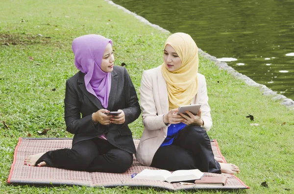 Muslim muda duduk di rumput hijau di taman dan membaca buku elektronik di ponsel mereka — Stok Foto