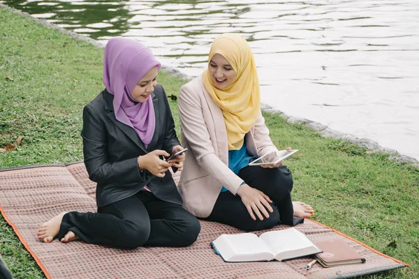 Potret wanita muda campuran yang menyenangkan duduk di rumput di taman musim panas yang hijau dan berdiskusi — Stok Foto