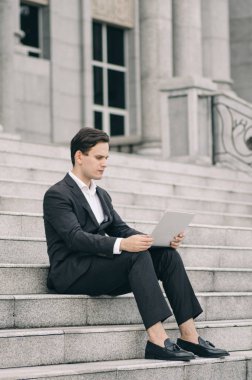 Kendine güvenen genç iş adamı merdivenlerde oturuyor ve laptopuyla çalışıyor. 