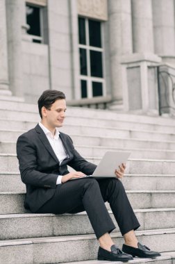 Gülümseyen genç iş adamı merdivenlerde oturuyor ve laptopuyla çalışıyor. 