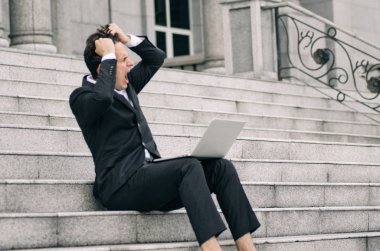 Hüsrana uğramış ve depresyonda olan genç bir iş adamı dizüstü bilgisayarıyla merdivenlerde oturuyor.