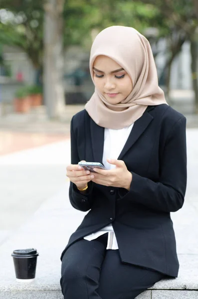 Retrato de hermosa joven mujer de negocios musulmana en traje negro sentado afuera y enviando mensajes de texto a su amigo usando el teléfono —  Fotos de Stock