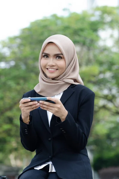 Retrato de la hermosa joven musulmana en traje negro y hijab sentado fuera usando el teléfono móvil —  Fotos de Stock