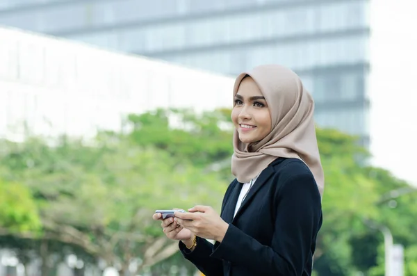 Обрезанный образ красивой молодой мусульманки на черном костюме и хиджабе, сидящих снаружи с помощью мобильного телефона — стоковое фото