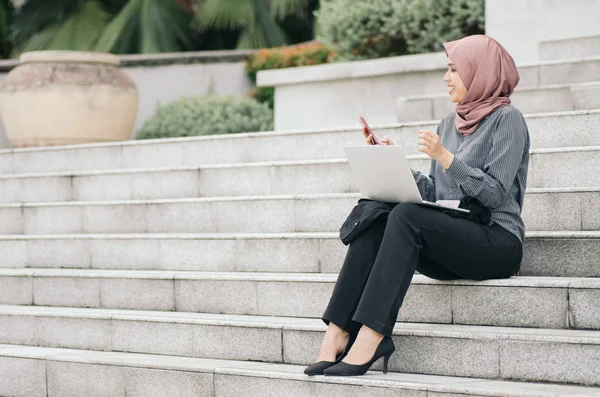 Elegant hijab businesswoman with laptop relaxing on stairs, smiling on her handphone