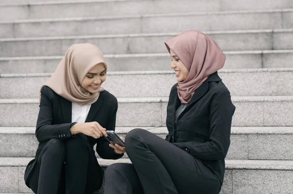 Glad ansikte, två unga företag hijab kvinna, sitter på trappor och diskuterar medan du håller mobiltelefon — Stockfoto