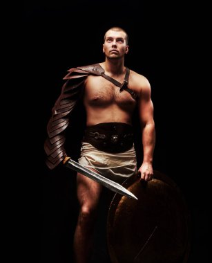 male gladiator with sword and armor on a black background looks into the camera. studio shot clipart